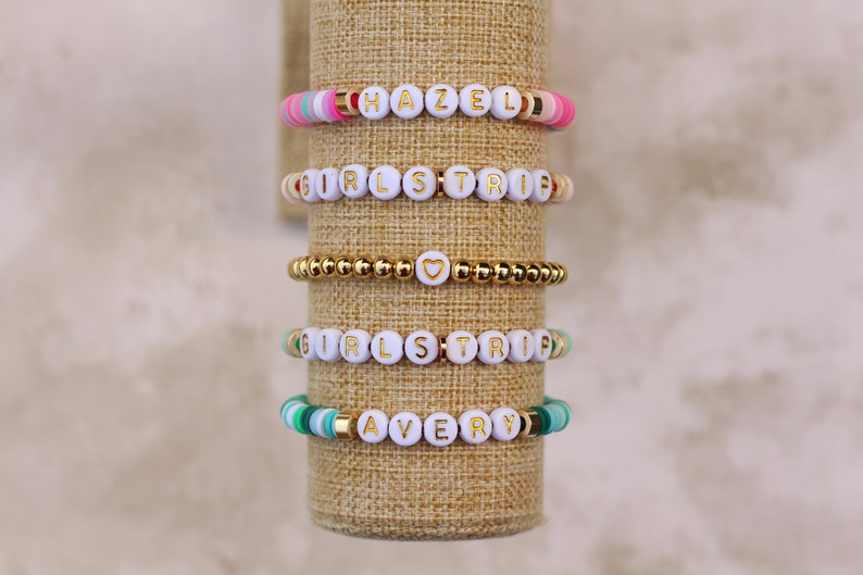 May include: Five beaded bracelets with white letter beads spelling "HAZEL", "GIRLS TRIP", and "AVERY". The bracelets feature gold and colorful beads in pink, green, and gold. One bracelet is all gold beads with a heart charm.