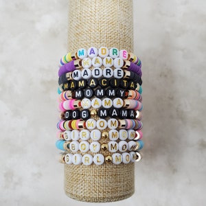 Pode incluir: Uma pilha de pulseiras de contas coloridas com pingentes de letras douradas que soletram palavras como "Mamacita", "Mommy", "Mama" e "Girl Mom".
