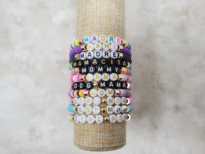 Pode incluir: Uma pilha de pulseiras de contas coloridas com pingentes de letras douradas que soletram palavras como "Mamacita", "Mommy", "Mama" e "Girl Mom".
