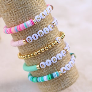 May include: A collection of colorful beaded bracelets with white letter beads spelling out words like "HAZE", "GIRLS", and "LOVE". The bracelets feature gold accents and are displayed on a tan fabric.