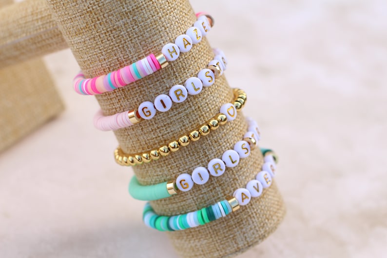 May include: A collection of colorful beaded bracelets with white letter beads spelling out words like "HAZE", "GIRLS", and "LOVE". The bracelets feature gold accents and are displayed on a tan fabric.