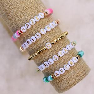 May include: Five beaded bracelets with white letter beads spelling out "HAZEL", "GIRLS TRIP", and "AVERY". The bracelets feature gold and colorful accent beads. The bracelets are displayed on a tan fabric.