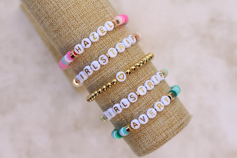 May include: Five beaded bracelets with white letter beads spelling out "HAZEL", "GIRLS TRIP", and "AVERY". The bracelets feature gold and colorful accent beads. The bracelets are displayed on a tan fabric.