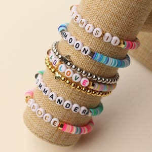 May include: A collection of colorful beaded bracelets with various words spelled out in white letter beads. The bracelets are displayed on a tan cylindrical stand. The bracelets feature gold and silver accents.