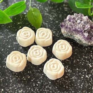 Könnte beinhalten: Sechs cremefarbene, rosenförmige Seifen auf einer dunklen, gesprenkelten Oberfläche. Grüne Blattranken und ein violetter Amethyst-Cluster befinden sich im Hintergrund. Die Seifen haben ein detailliertes Blumendesign.