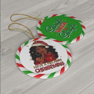 Puede incluir: Dos adornos navide&ntilde;os redondos. Un adorno presenta una ilustraci&oacute;n de dibujos animados de una mujer negra con un gorro de Pap&aacute; Noel y un su&eacute;ter rojo con el texto "Have a Melanin Christmas". El otro adorno es verde con rayas blancas y el texto "Sleigh Queen Sleigh".