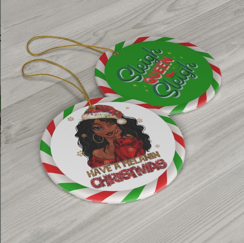 Puede incluir: Dos adornos navide&ntilde;os redondos. Un adorno presenta una ilustraci&oacute;n de dibujos animados de una mujer negra con un gorro de Pap&aacute; Noel y un su&eacute;ter rojo con el texto "Have a Melanin Christmas". El otro adorno es verde con rayas blancas y el texto "Sleigh Queen Sleigh".