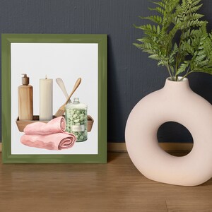 Op de afbeelding: Een ingelijste illustratie van spa-artikelen, waaronder een kaars, zeep, handdoeken en een pot badzout. De illustratie is in een groene lijst en ligt op een houten oppervlak.