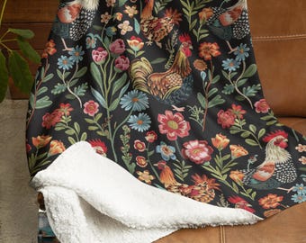 May include: A black throw blanket with a floral and rooster pattern. The blanket has a white sherpa lining and is draped over a brown leather sofa. The design includes colourful flowers and detailed rooster illustrations.