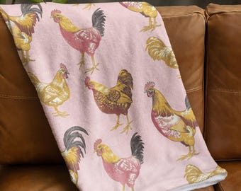 May include: A pink blanket with a repeating pattern of roosters in yellow, red, and brown. The blanket is draped over a brown leather sofa, highlighting the soft texture and the detailed rooster illustrations. The blanket is made of a soft material.