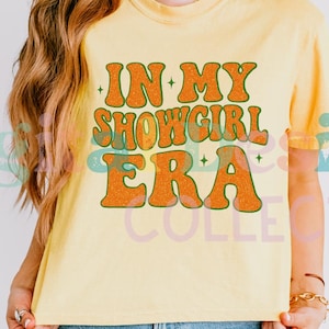 May include: Pale yellow t-shirt featuring the phrase "IN MY SHOWGIRL ERA" in orange, glittery letters with a green outline. The short-sleeved top is a light yellow colour.