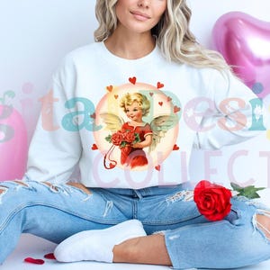 Könnte beinhalten: Ein weißer Pullover mit einer Vintage-Illustration eines Amor-Engels, der einen Strauß roter Rosen hält. Das Design ist von roten Herzen umgeben.