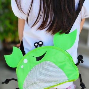 May include: A green crab-shaped backpack with a white mesh front and black accents. The backpack has two large green claw-like flaps on the sides and a black strap. The backpack is worn by a person wearing a white t-shirt.