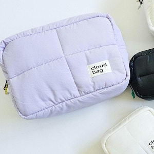 Op de afbeelding: Een lavendelkleurige, gewatteerde cloud bag met een rechthoekige vorm en een ritssluiting. De tas heeft een gestikt rasterpatroon en een klein wit label met de tekst "cloud bag". Andere tassen in zwart en wit zijn ook zichtbaar.