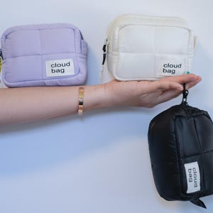 Może przedstawiać: Trzy pikowane torby w kształcie chmury w różnych kolorach: lawendowym, białym i czarnym. Każda torba ma kwadratowy kształt z zapięciem na zamek błyskawiczny i małą metką z napisem "cloud bag". Torby są trzymane w dłoni.