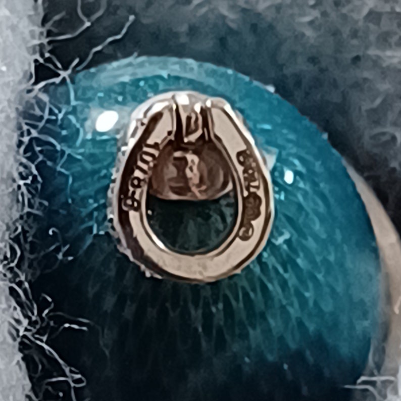 Puede incluir: Primer plano de una cuenta de vidrio azul turquesa con un bucle de metal plateado. El bucle est&aacute; grabado con el texto "925" y "925" a cada lado.