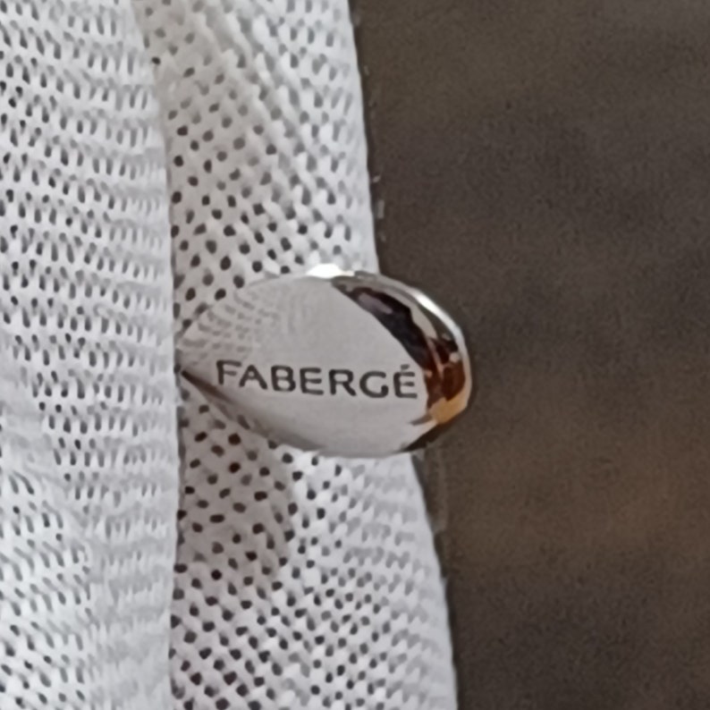Puede incluir: Una etiqueta ovalada de color plateado con la palabra "FABERG&Eacute;" en relieve.