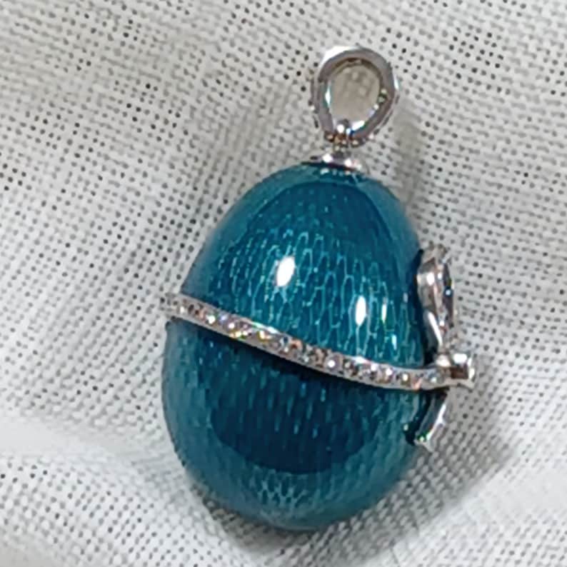 Puede incluir: Pendiente en forma de huevo azul turquesa con una banda plateada y un lazo plateado en la parte superior. El pendiente tiene una superficie texturizada.