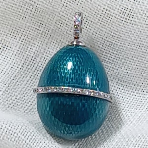 Puede incluir: Un colgante de huevo de esmalte azul turquesa con una banda de piedras brillantes transparentes alrededor del centro. El colgante tiene un bucle plateado en la parte superior para colgar.