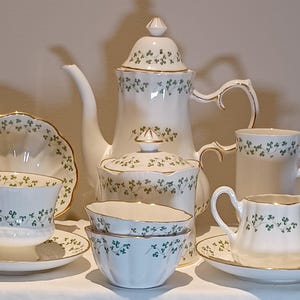 May include: A white porcelain tea set with a green shamrock pattern. The set includes a teapot, sugar bowl, creamer, cups, saucers, and bowls. Gold trim accents the edges of the pieces. A classic and elegant design.