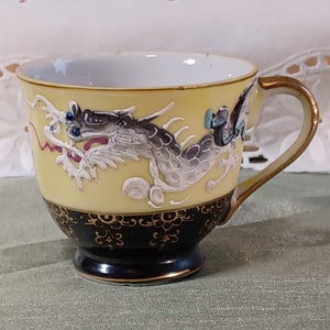 May include: A yellow teacup with a black base and a gold handle. The cup features a detailed, raised relief of a dragon in shades of gray, white, and blue. The rim and handle are trimmed with gold, and the base has a black band with gold floral designs.