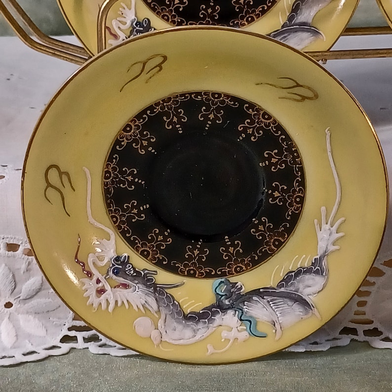 May include: A set of yellow and black porcelain saucers with gold trim. Each saucer features a detailed dragon design in white, gray, and blue, with a black center and gold floral accents. The saucers are part of a tea set.