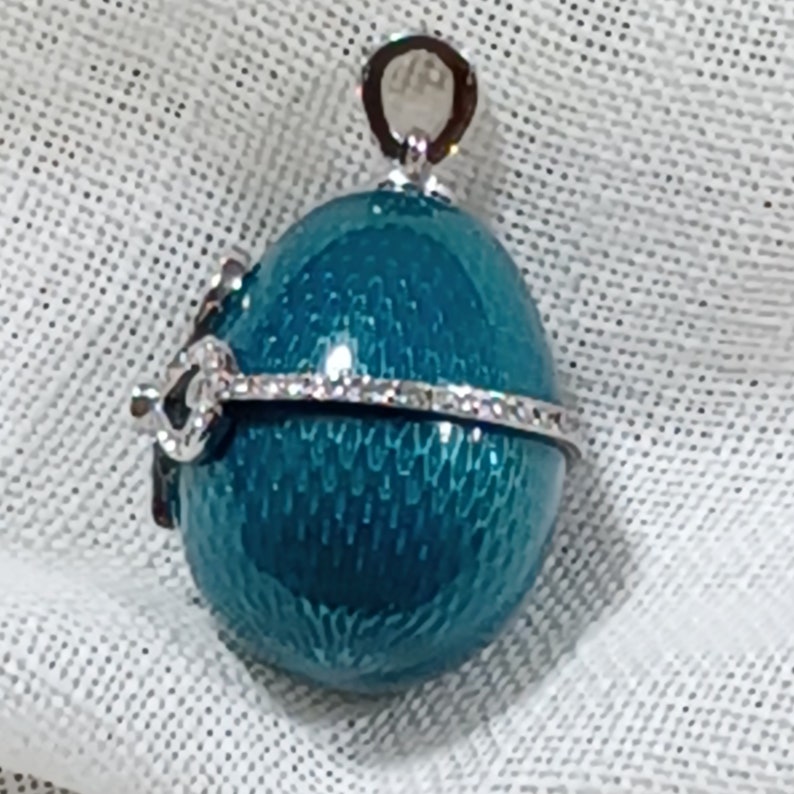 Puede incluir: Pendiente en forma de huevo azul turquesa con una banda plateada de diamantes de imitaci&oacute;n alrededor del centro. El pendiente tiene un bucle plateado en la parte superior para sujetar una cadena.