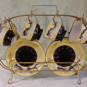 May include: A set of six teacups and saucers with a gold-colored metal rack. The cups and saucers are yellow with black and gold accents, and feature a dragon design. The cups are suspended from the rack.