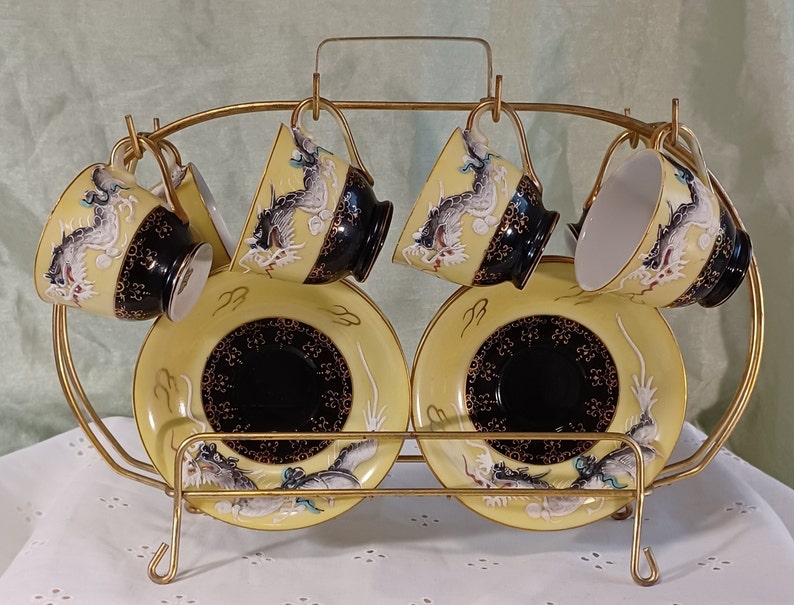 May include: A set of six teacups and saucers with a gold-colored metal rack. The cups and saucers are yellow with black and gold accents, and feature a dragon design. The cups are suspended from the rack.