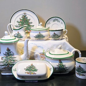 May include: A collection of white ceramic Christmas-themed tableware. The set includes a teapot, plates, cups, a sugar bowl, a creamer, and a butter dish. Each piece features a green Christmas tree design with colorful ornaments and a green trim.