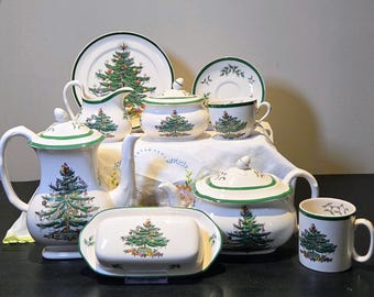 May include: A collection of white ceramic Christmas-themed tableware. The set includes a teapot, plates, cups, a sugar bowl, a creamer, and a butter dish. Each piece features a green Christmas tree design with colorful ornaments and a green trim.