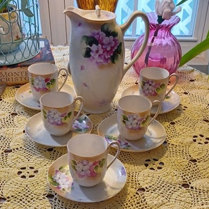 May include: A vintage porcelain tea set featuring a cream-colored teapot and six matching teacups and saucers. The set is adorned with hand-painted purple and white floral designs. The teacups and teapot have gold trim. The set is displayed on a yellow lace tablecloth.