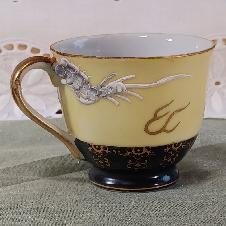 May include: A yellow teacup with a gold handle and rim, featuring a white and gray dragon design. The base is black with a gold floral pattern. The teacup is a delicate piece of Asian-inspired tableware.