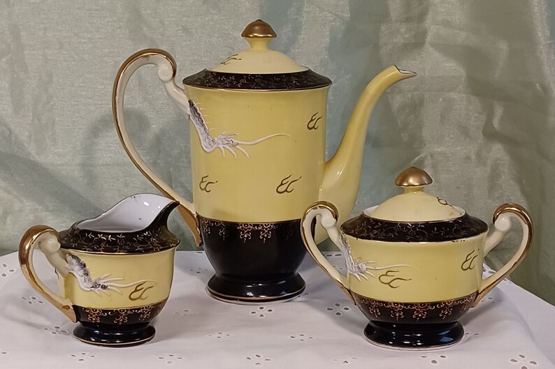 May include: A vintage tea set with a yellow and black design. The set includes a teapot, creamer, and sugar bowl. Each piece features a dragon motif and gold accents. The teapot has a tall, slender spout and a matching lid.