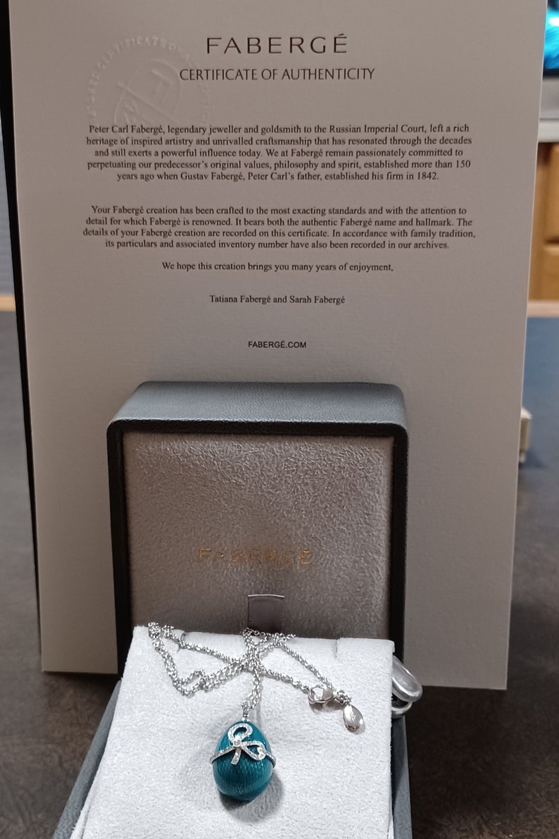 Puede incluir: Un collar con un colgante de huevo Faberg&eacute; azul turquesa, una cadena de plata y un lazo engastado con diamantes. El collar se muestra en una caja negra con el logotipo de Faberg&eacute;.