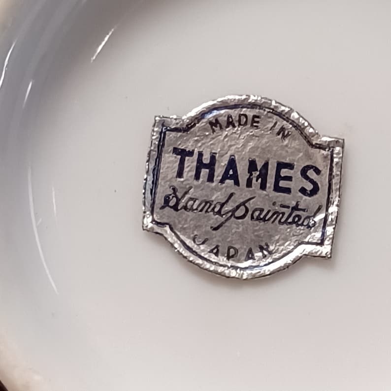 May include: A close-up of a white ceramic surface with a silver label. The label reads "MADE IN THAMES Hand Painted JAPAN". The label has a silver border and black text.