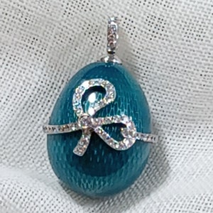 Puede incluir: Un colgante en forma de huevo azul turquesa con un dise&ntilde;o de lazo plateado incrustado con diamantes de imitaci&oacute;n transparentes.