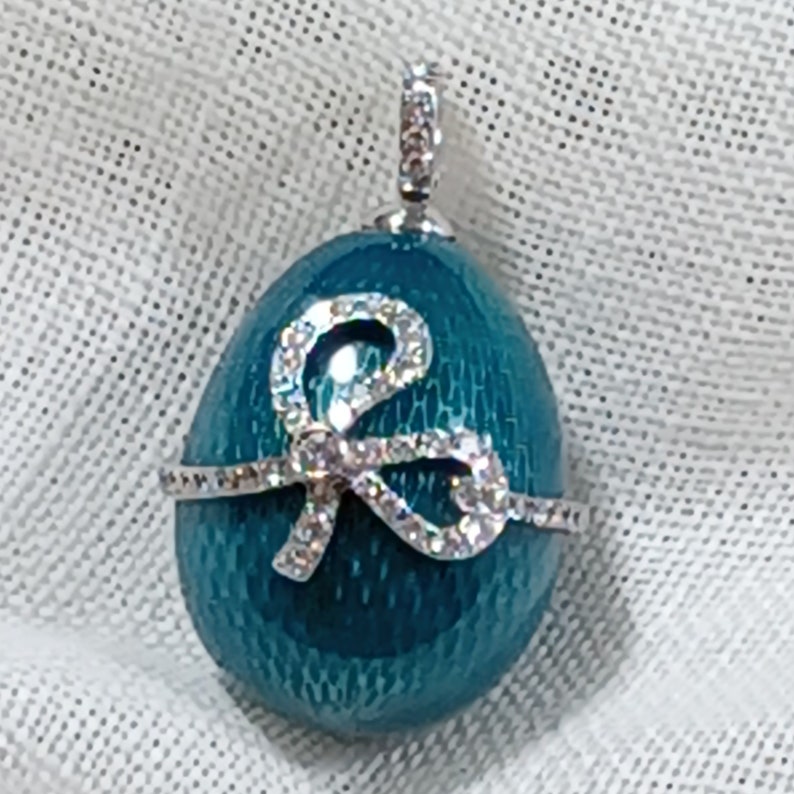 Puede incluir: Un colgante en forma de huevo azul turquesa con un dise&ntilde;o de lazo plateado incrustado con diamantes de imitaci&oacute;n transparentes.