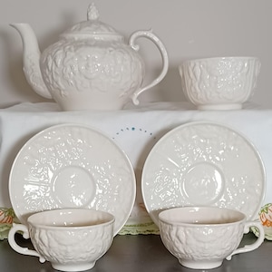 May include: A white ceramic tea set featuring a teapot, two teacups, two saucers, and a small bowl. The set has a textured floral design. The teapot has a lid with a decorative finial. The teacups have handles.