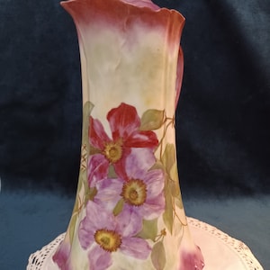 May include: A tall, antique porcelain pitcher with a lid. The pitcher is painted with vibrant red and purple flowers and green leaves. The rim and base are a deep burgundy color, and the handle and lid have gold accents. The pitcher is on a white lace doily.