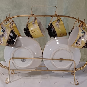 May include: A vintage tea set with six cups and saucers, displayed on a gold-colored metal rack. The cups feature a yellow background with a black rim and a dragon design. The saucers are white. A decorative and functional item.