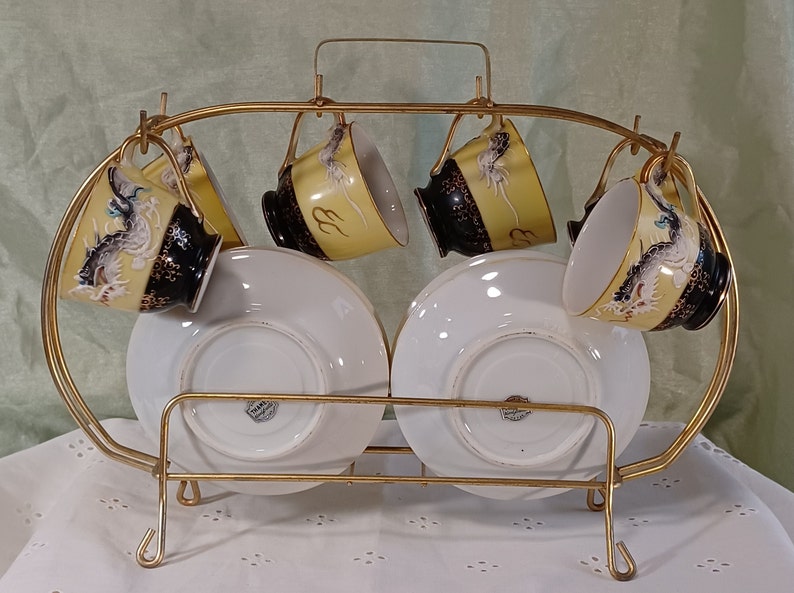 May include: A vintage tea set with six cups and saucers, displayed on a gold-colored metal rack. The cups feature a yellow background with a black rim and a dragon design. The saucers are white. A decorative and functional item.