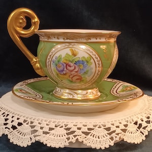 May include: A vintage teacup and saucer set. The cup and saucer are green with gold trim and floral designs. The cup has a decorative gold handle. The set is displayed on a white lace doily.