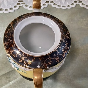 May include: Overhead view of a vintage sugar bowl. The bowl is yellow with a black rim featuring gold floral designs. It has two gold handles and a white interior. The bowl is on a light green surface.
