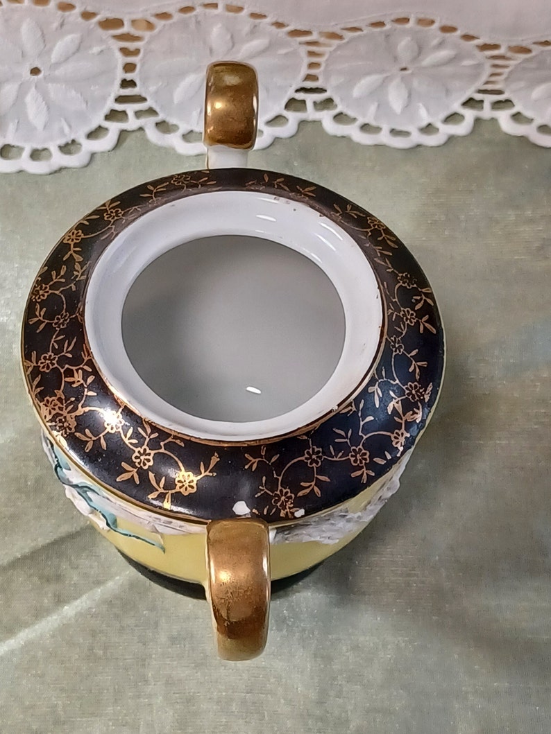 May include: Overhead view of a vintage sugar bowl. The bowl is yellow with a black rim featuring gold floral designs. It has two gold handles and a white interior. The bowl is on a light green surface.