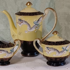 May include: A yellow and black porcelain tea set with a dragon motif. The set includes a teapot, sugar bowl, and creamer. The teapot has a tall, curved spout and a gold-colored handle. The dragon is white and gray with blue accents.