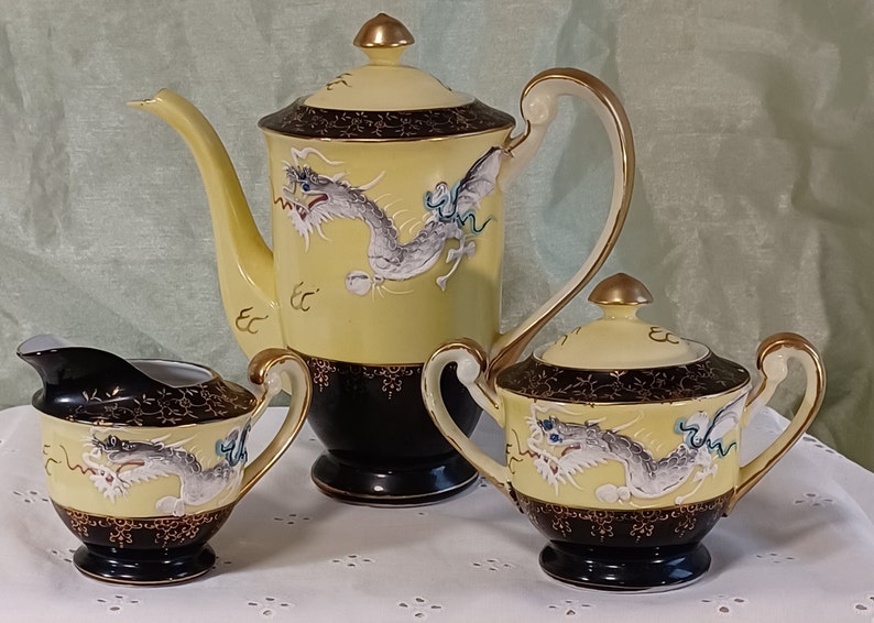 May include: A yellow and black porcelain tea set with a dragon motif. The set includes a teapot, sugar bowl, and creamer. The teapot has a tall, curved spout and a gold-colored handle. The dragon is white and gray with blue accents.