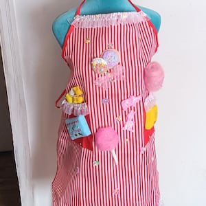 May include: A red and white striped apron with a variety of candy and sweet treats attached. The apron has a pocket with the words "Candy and Sweet Shop" embroidered on it. The apron is on a white mannequin with a pink cotton candy and popcorn headband.