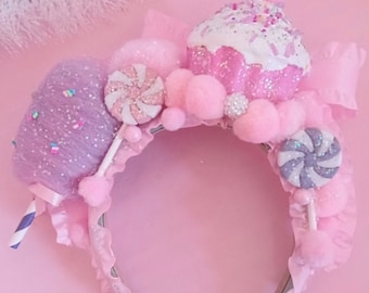 Könnte beinhalten: Ein rosa und lilafarbenes Stirnband mit einem Cupcake, Zuckerwatte und Lutschern. Das Stirnband ist mit weißen Pompons und Glitzer verziert.