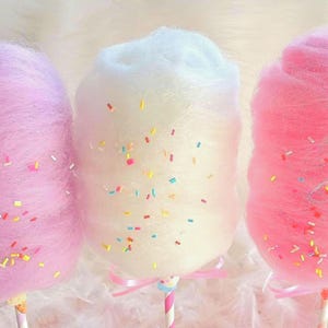Puede incluir: Tres coloridos algodones de azúcar en palitos. El algodón de azúcar es rosa, blanco y morado, y cada uno está rociado con chispas de colores. Los dulces están atados con cintas rosas.
